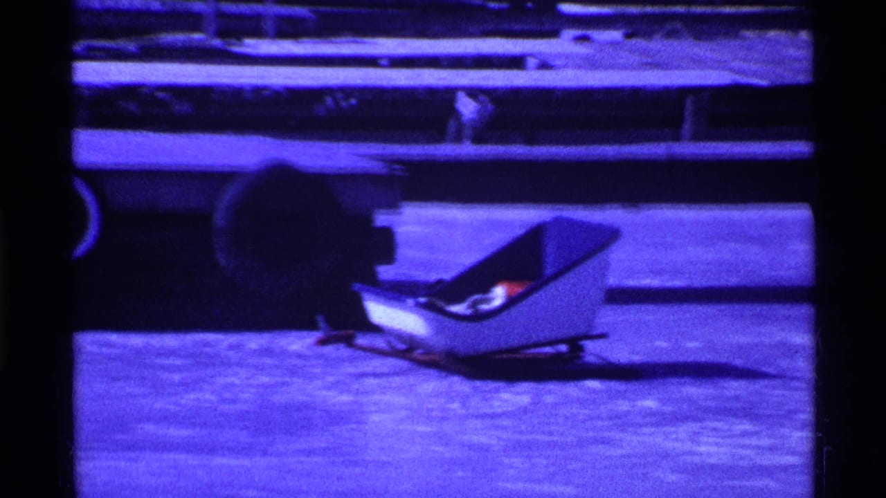 Blue-tinted 1969 Ohio home-movie frame with a low sled-like vehicle on ice or packed snow beside horizontal railings.