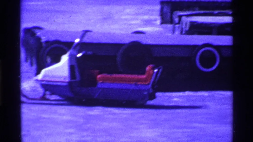 Blue-tinted 1969 Ohio 8mm frame with a snowmobile-like vehicle and red seat beside a large dark vehicle on frozen ground.