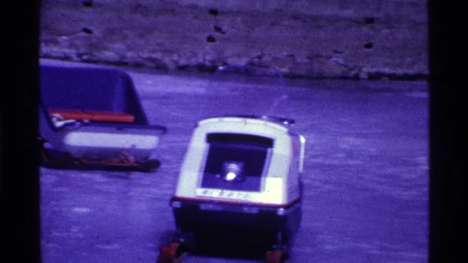 1969 Ohio home-movie frame with a small white-and-blue toy-like vehicle on pavement near the lakeside.