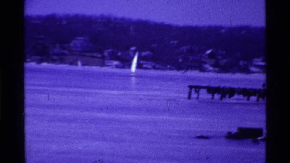 Purple-toned 8mm frame of an iceboat sail crossing a frozen Ohio lake, with trees and shoreline structures in the distance.