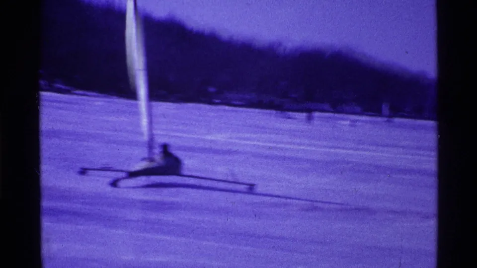 Closer 1969 Ohio home-movie frame showing an adult seated on a small iceboat with runners and a tall white sail on the ice.