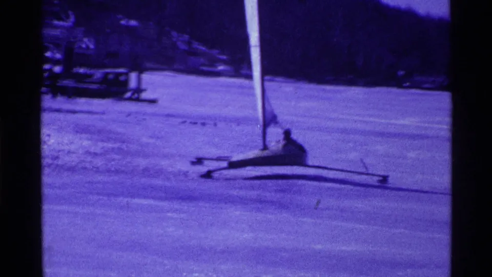Vintage 8mm frame from Ohio showing an adult operating a small iceboat across the frozen lake, with shoreline and trees behind.