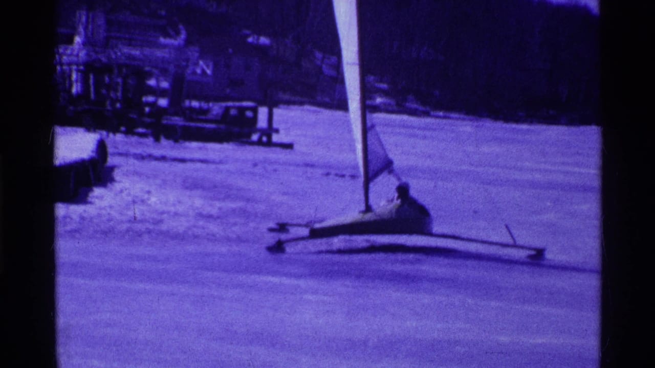 Small iceboat with a seated sailor crossing a frozen lake near a snowy shoreline.