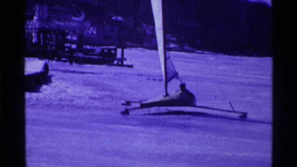 1969 Ohio 8mm frame of a small iceboat moving across a frozen lake, with a seated sailor and distant lakeside structures.