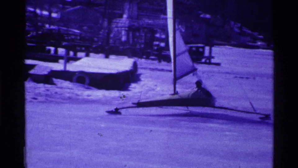 Small sail iceboat on a frozen Ohio lake in 1969, seen in purple-tinted 8mm footage with shoreline structures behind it.