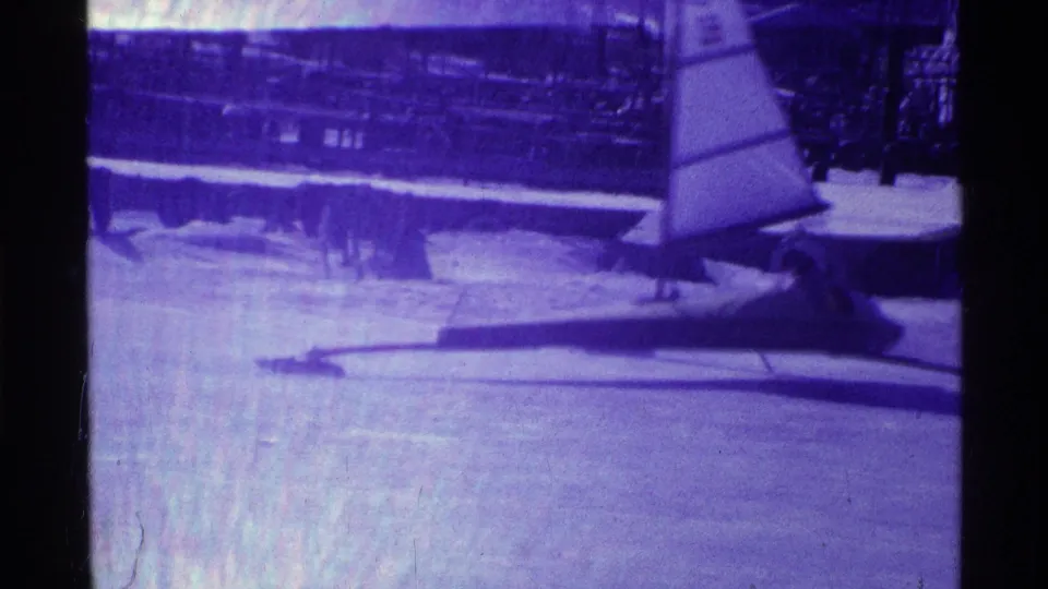 Purple-tinted 8mm frame showing the iceboat passing closer to camera on a frozen Ohio lake, with sail and runner visible.