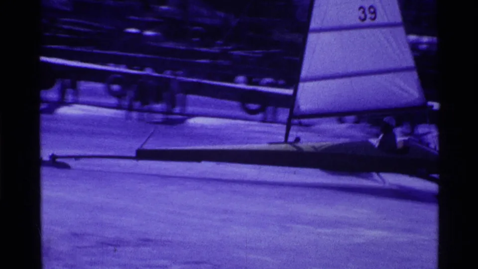 1969 Ohio home-movie frame of an iceboat with sail number 39, a seated sailor, and dark shoreline structures behind it.
