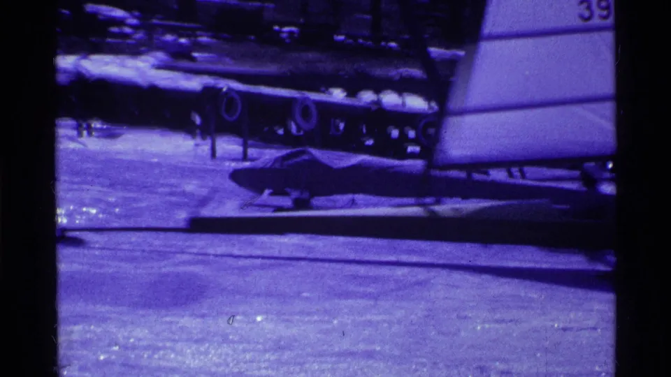 1969 Ohio home-movie frame of a sail-equipped ice craft on a frozen lake with distant dark shapes behind it.