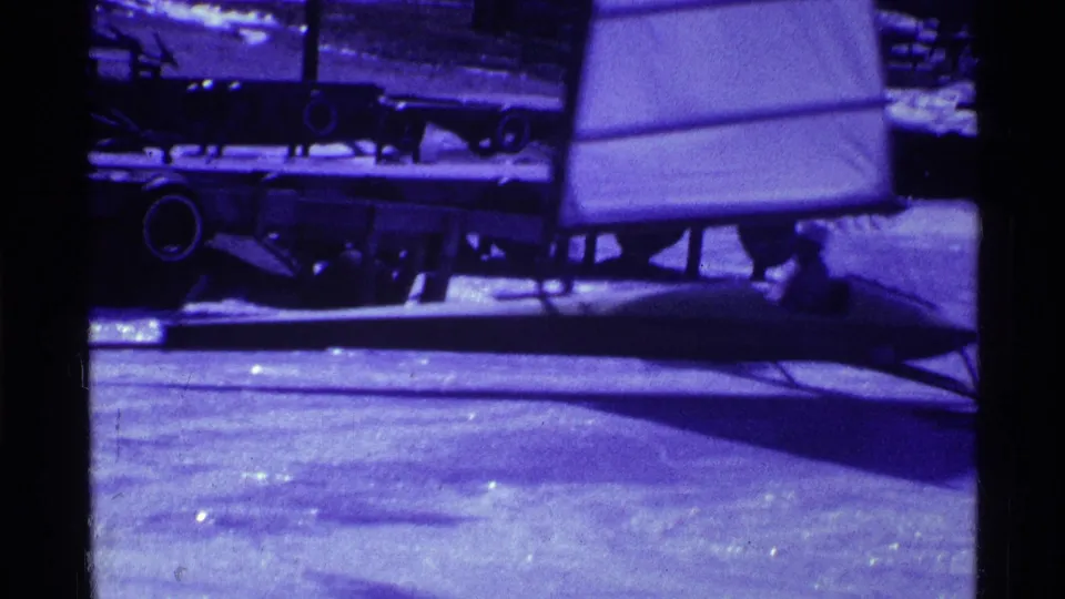 1969 Ohio frame showing a person in dark winter clothing seated near a low ice-sailing craft on the frozen lake.