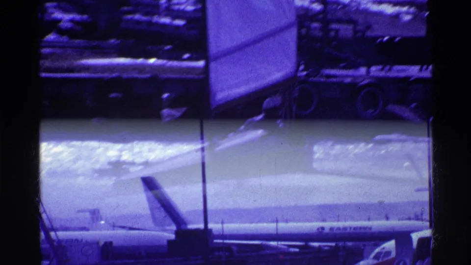 1969 Ohio airport home-movie frame showing a passenger jet on the tarmac through window framing, with airport structures and a purple color cast.