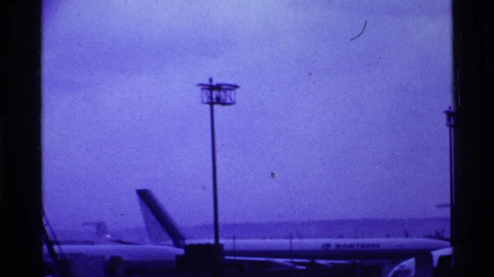 1969 Ohio airport frame with a jet parked or taxiing beyond the window, a tall light pole centered against the hazy sky.