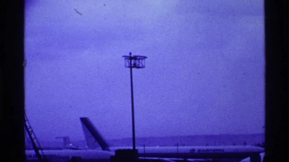 1969 Ohio airport home-movie still showing a light pole, aircraft tail, and low terminal-side details through tinted, grainy 8mm footage.