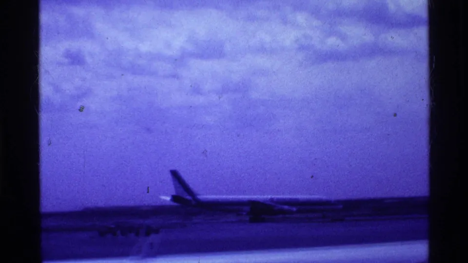 1969 Ohio airport footage frame with a passenger aircraft silhouette low across the runway, tinted blue-purple by the film scan.