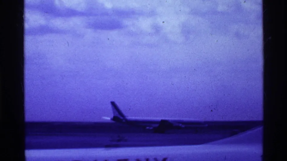 1969 Ohio airport frame showing a distant runway aircraft and a large foreground aircraft surface, likely filmed from a traveler viewpoint.