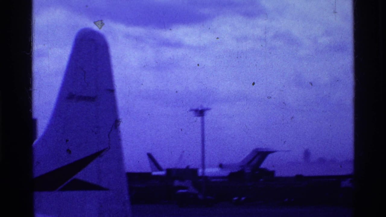 1969 Ohio airport view with a parked aircraft tail in the foreground and runway activity beyond under a cloudy blue-cast sky.