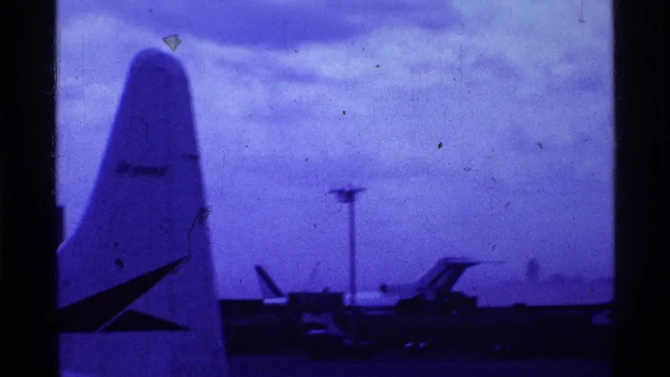 1969 Ohio airport view with a parked aircraft tail in the foreground and runway activity beyond under a cloudy blue-cast sky.