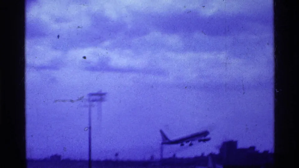 1969 Ohio home-movie frame showing an airliner climbing away from the airport runway into a cloudy, blue-tinted sky.