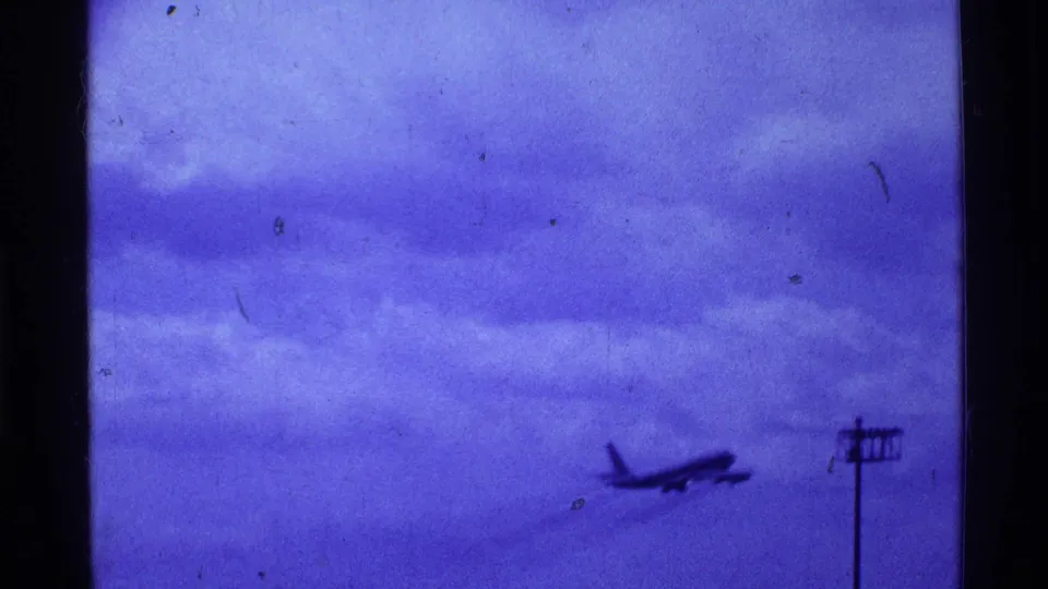 1969 Ohio home-movie frame of a jet airliner ascending across a tinted sky, with a utility pole near the airport edge.