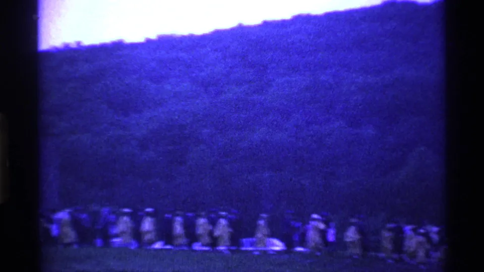 In 1969 Ohio 8mm footage, a large group in light robes or uniforms appears in formation across a field under a wooded hillside.