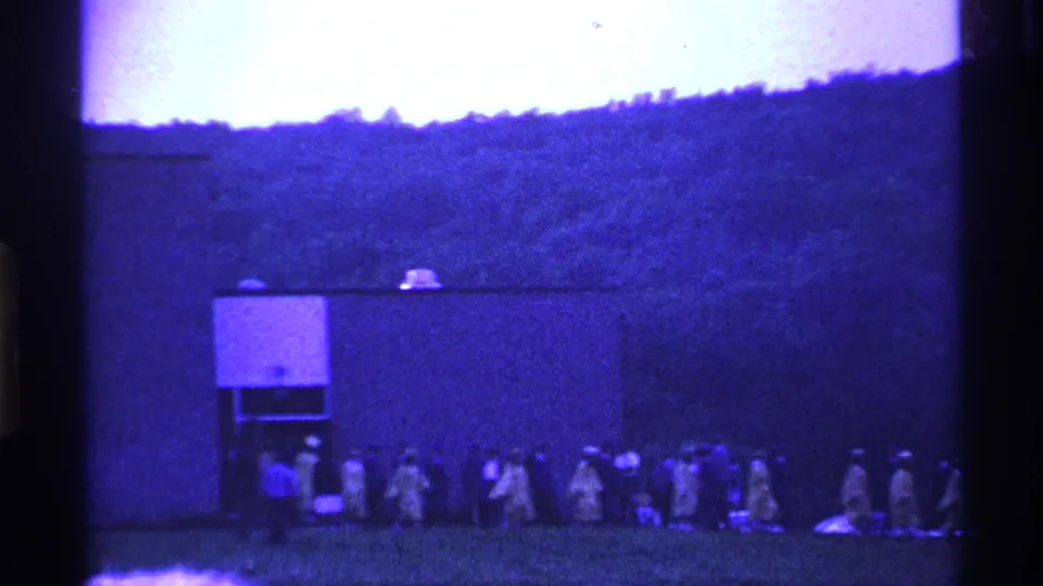 A distant marching line passes a small camp-like structure in Ohio home-movie footage from 1969, with a wooded slope in the background.