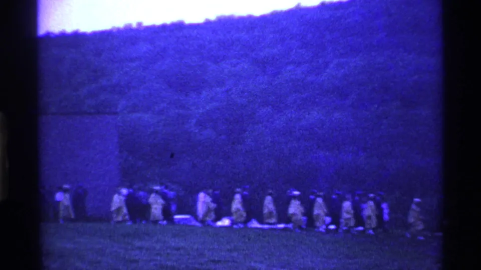 The 1969 Ohio scene shows a distant row of people walking in formation across a grassy area, filmed with soft blue 8mm texture.