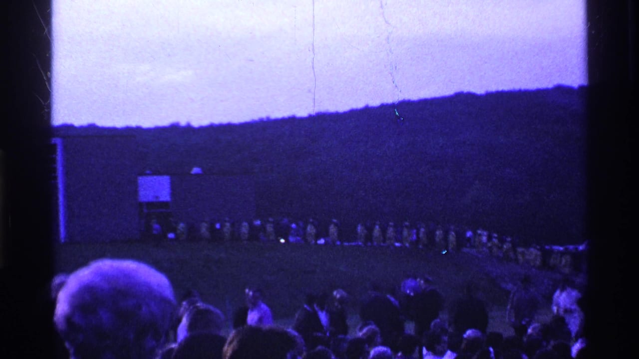 Crowd watches a long line of people crossing a grassy field below a wooded hill.