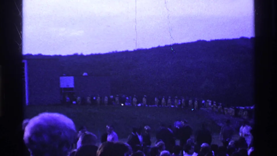 1969 Ohio 8mm frame showing spectators in the foreground and a distant line of people in light raincoats across a field.