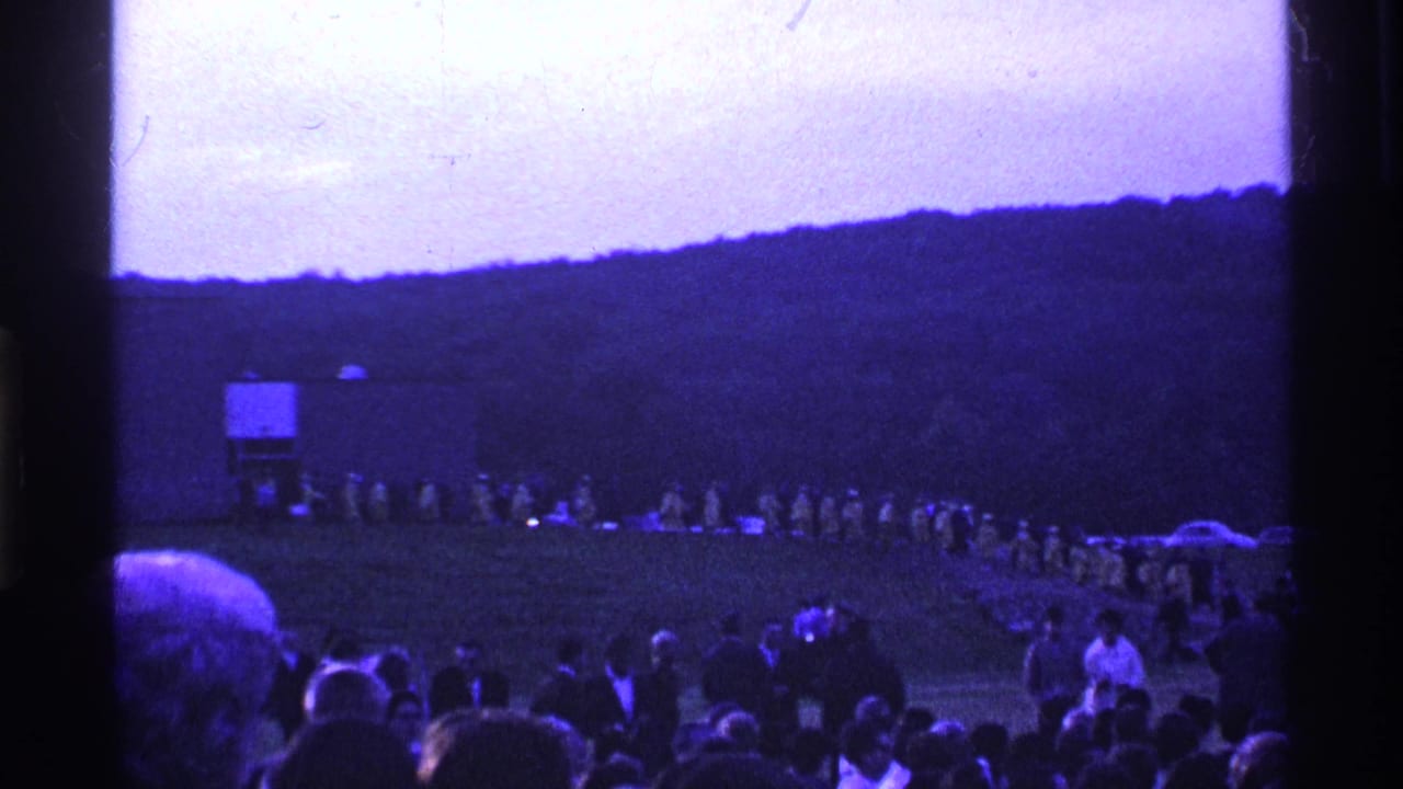 Large outdoor crowd faces a curved line of people near a field structure and hillside.