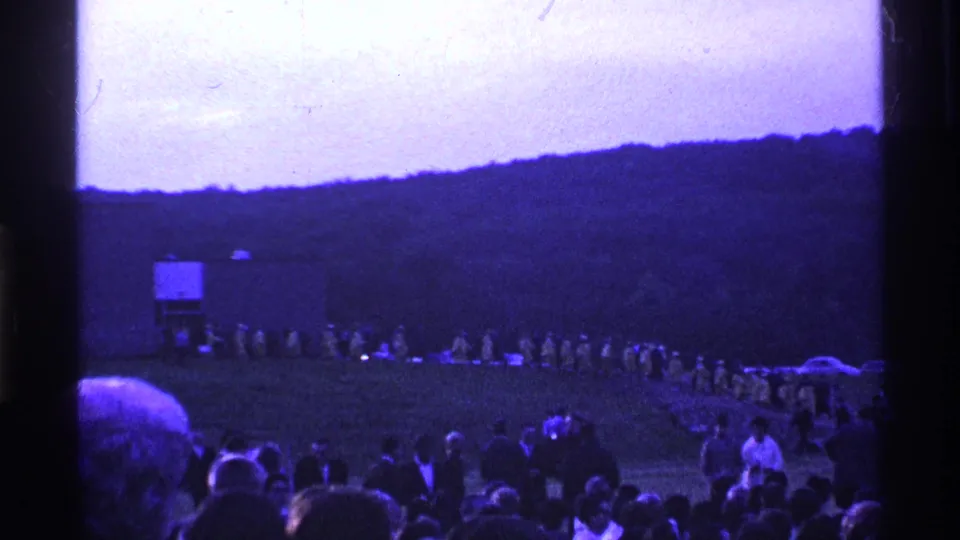 1969 Ohio home-movie frame with a foreground crowd watching a curved procession across grass near a simple structure and wooded hill.