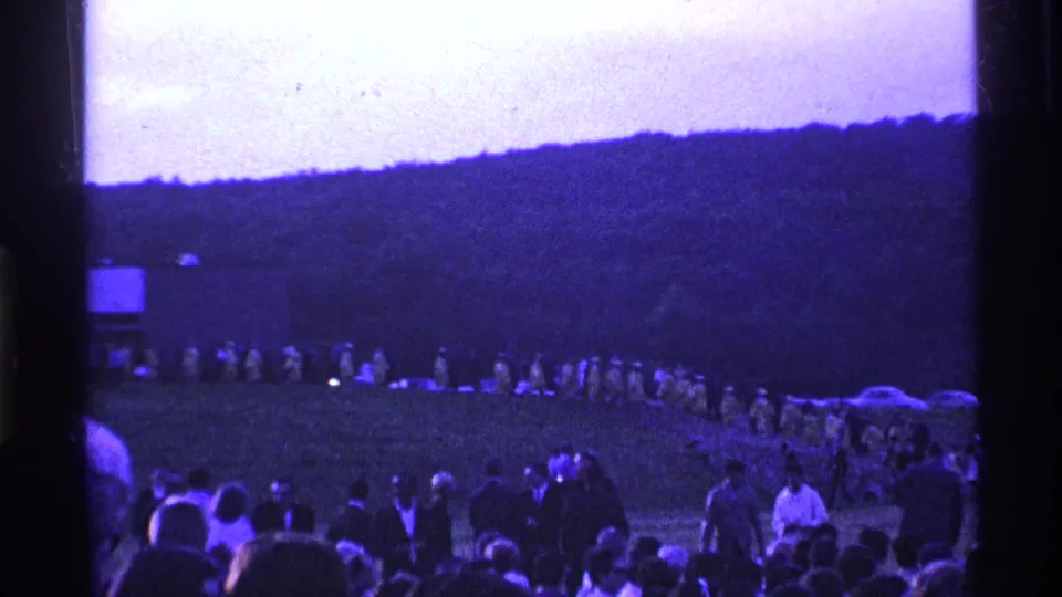 1969 Ohio 8mm frame showing a distant raincoat line across the field, foreground spectators, parked cars, and a dark wooded hillside.
