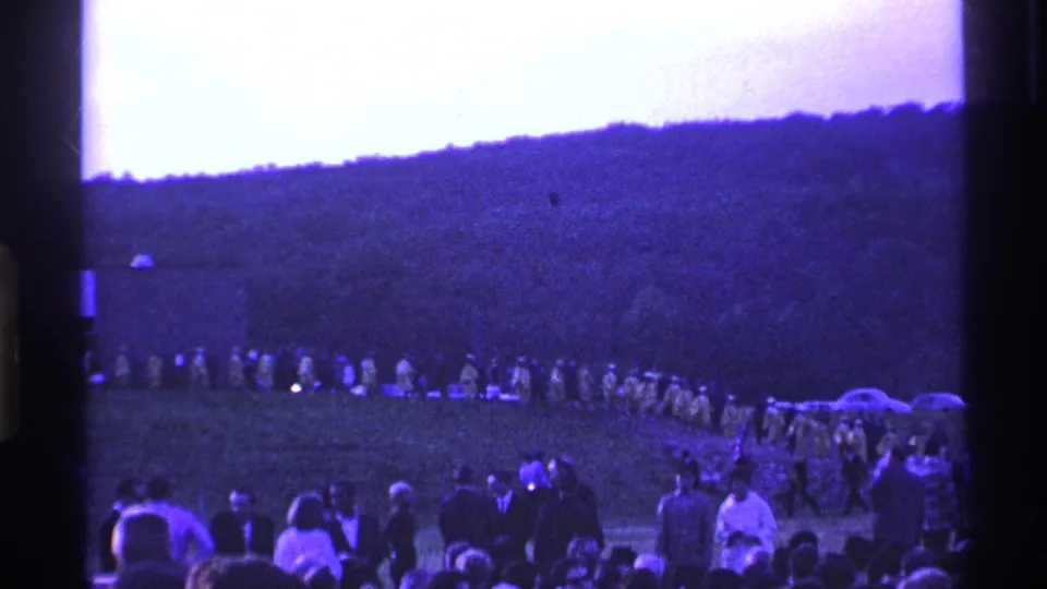 1969 Ohio home-movie frame with spectators in front, a distant procession on grass, parked cars, and a wooded hill under shifted color.