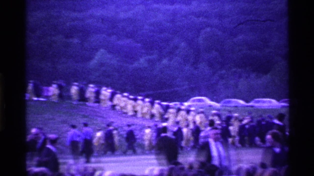 1969 Ohio home-movie frame showing yellow-robed graduates processing across grass with spectators and parked cars behind them.