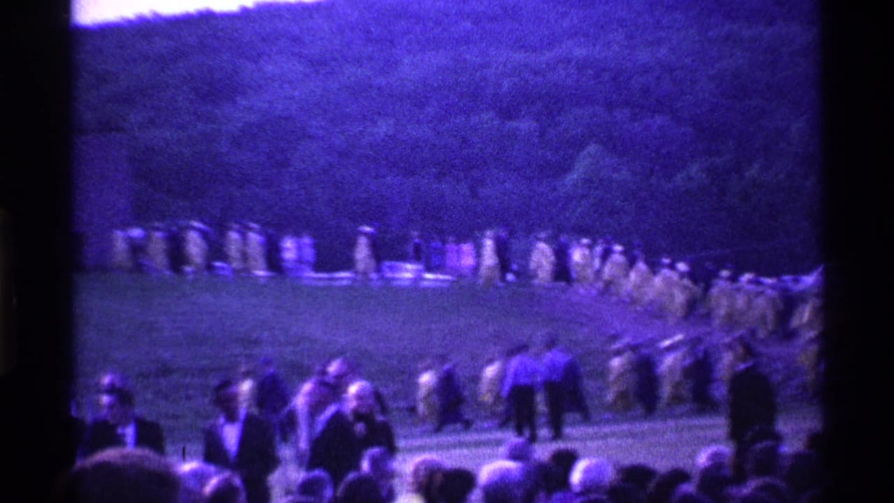 Yellow-robed graduates appear in procession across a grassy Ohio hillside, with a crowd and parked cars visible in the distance.