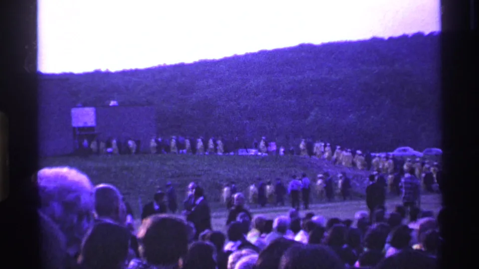 Wide 8mm view of an outdoor 1969 Ohio graduation, with audience members below and yellow-robed graduates along the hill.
