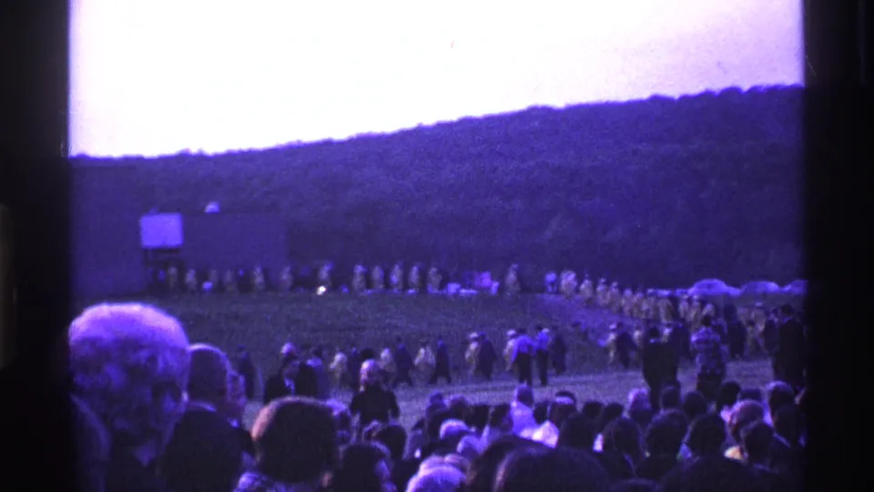 Outdoor graduation scene in Ohio, 1969, with seated and standing spectators watching a distant line of yellow-robed graduates.