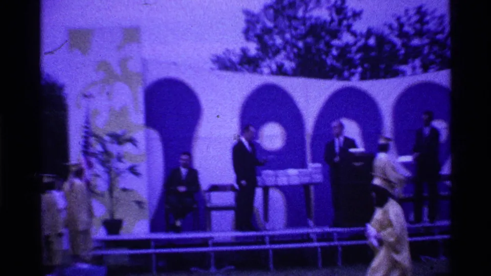 1969 Ohio graduation footage showing a graduate crossing an outdoor stage toward suited officials near a diploma table and arched backdrop.