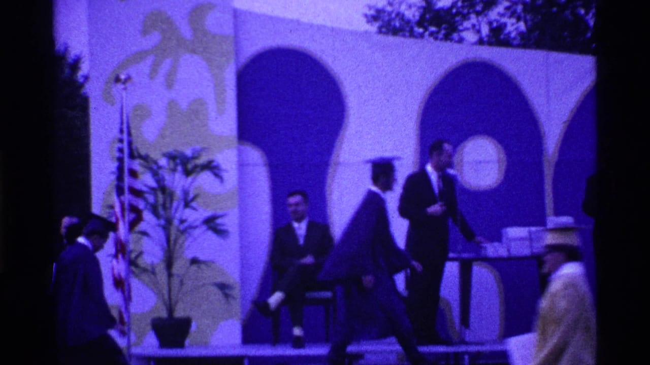 Graduate in dark gown walks near officials on a decorated outdoor stage with arches and a potted plant.