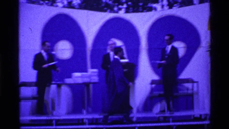 During the Ohio graduation ceremony, a robed graduate stands near suited officials and a table stacked with diploma boxes or papers.
