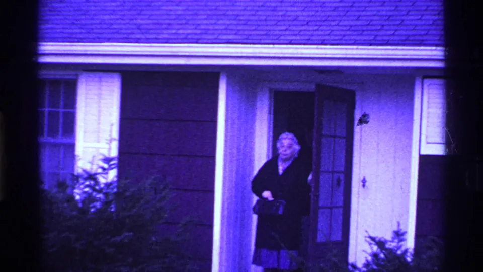 An older woman in a dark coat stands at the open front door of an Ohio house in purple-shifted 1969 8mm footage.