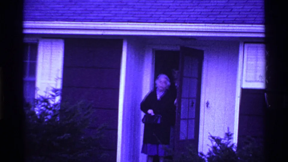The older woman stands near the open doorway of a suburban Ohio house, with shrubs and siding visible in the purple 8mm frame.