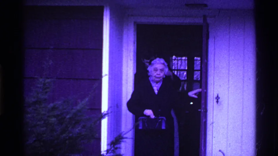 Closer 1969 Ohio home-movie frame of an older woman with white hair and a handbag at a house entrance.
