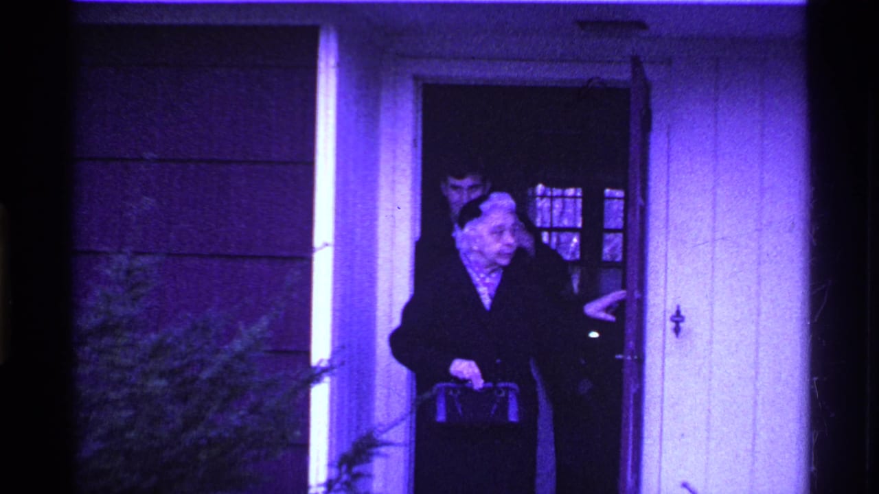 Elderly woman in a dark coat stands at a front doorway with a younger man behind her.
