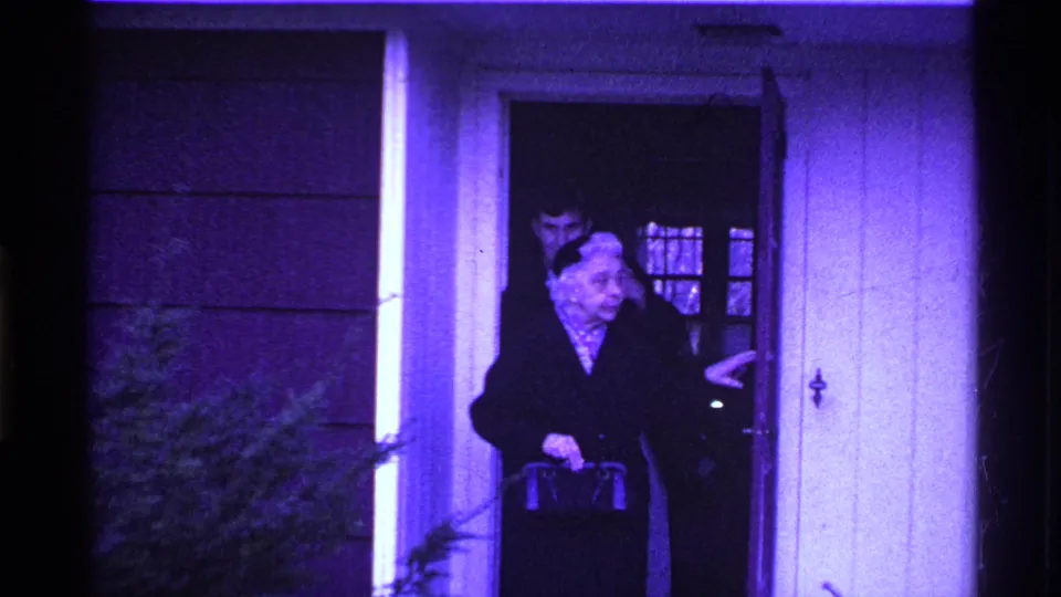 In metadata-supported 1969 Ohio home-movie footage, an elderly woman steps from a house doorway while a younger man stands behind her.