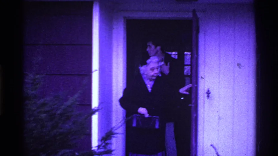 The elderly woman pauses outside the open door with her handbag, while the younger man remains partly visible in the doorway.