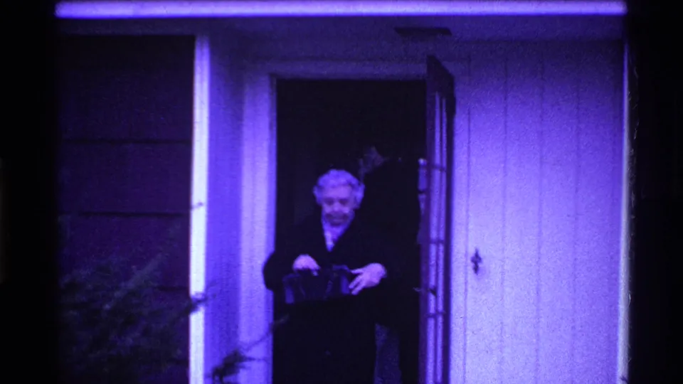 The woman smiles toward the camera outside the front door, holding or adjusting a small handbag in the purple-tinted frame.