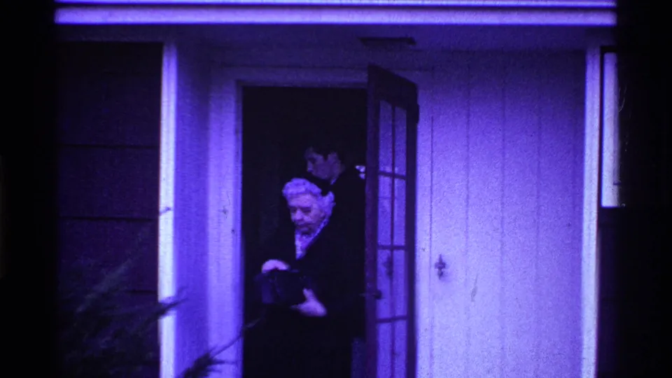 At the end of the clip, the elderly woman stands near the open door with the younger man still visible behind her in shadow.
