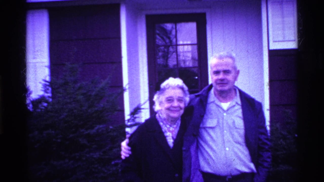 Older woman and man stand arm in arm outside a house doorway.