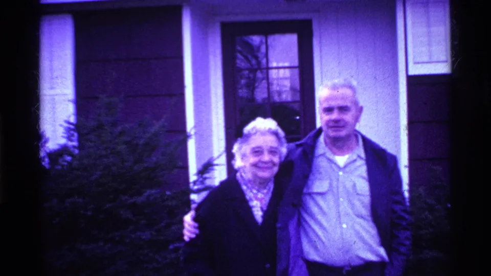 In Ohio 1969 home-movie footage, an older couple stands close together outside a house with windows and shrubs behind them.