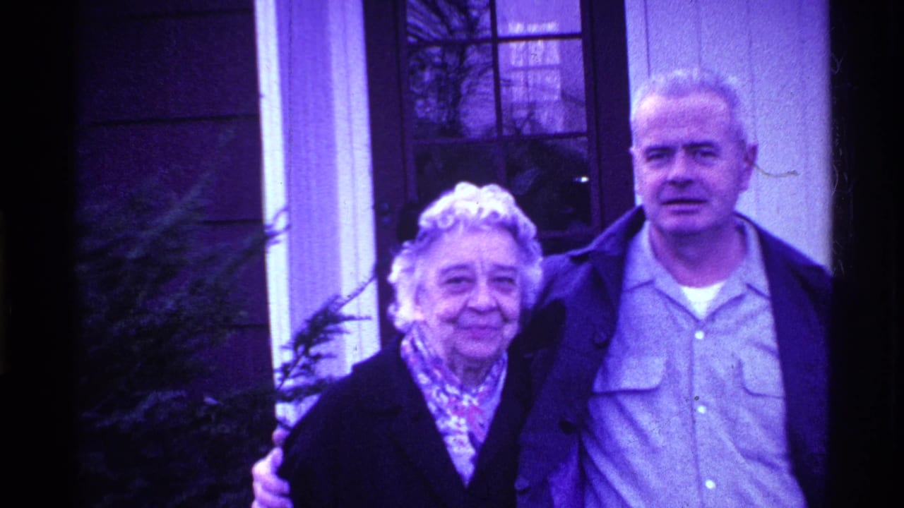 Older couple stands close together near a house window and doorway.