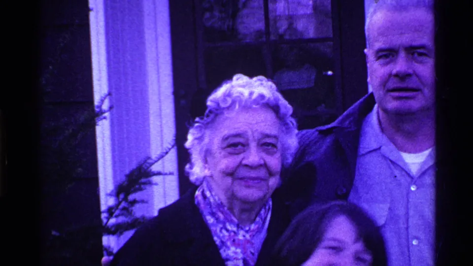 The couple stands outside the home while a younger-looking person is partially visible at the bottom edge of the frame.